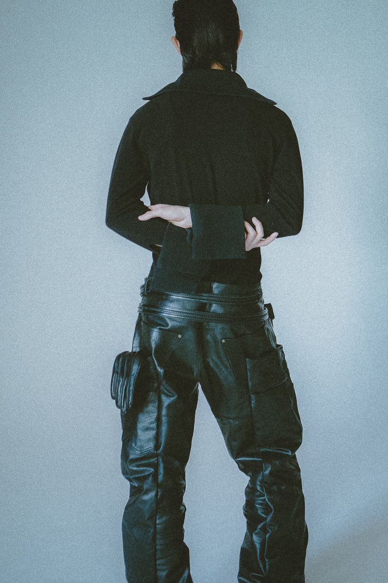 Back view of a person in a black jacket and leather pants standing in a light studio with their arms crossed.