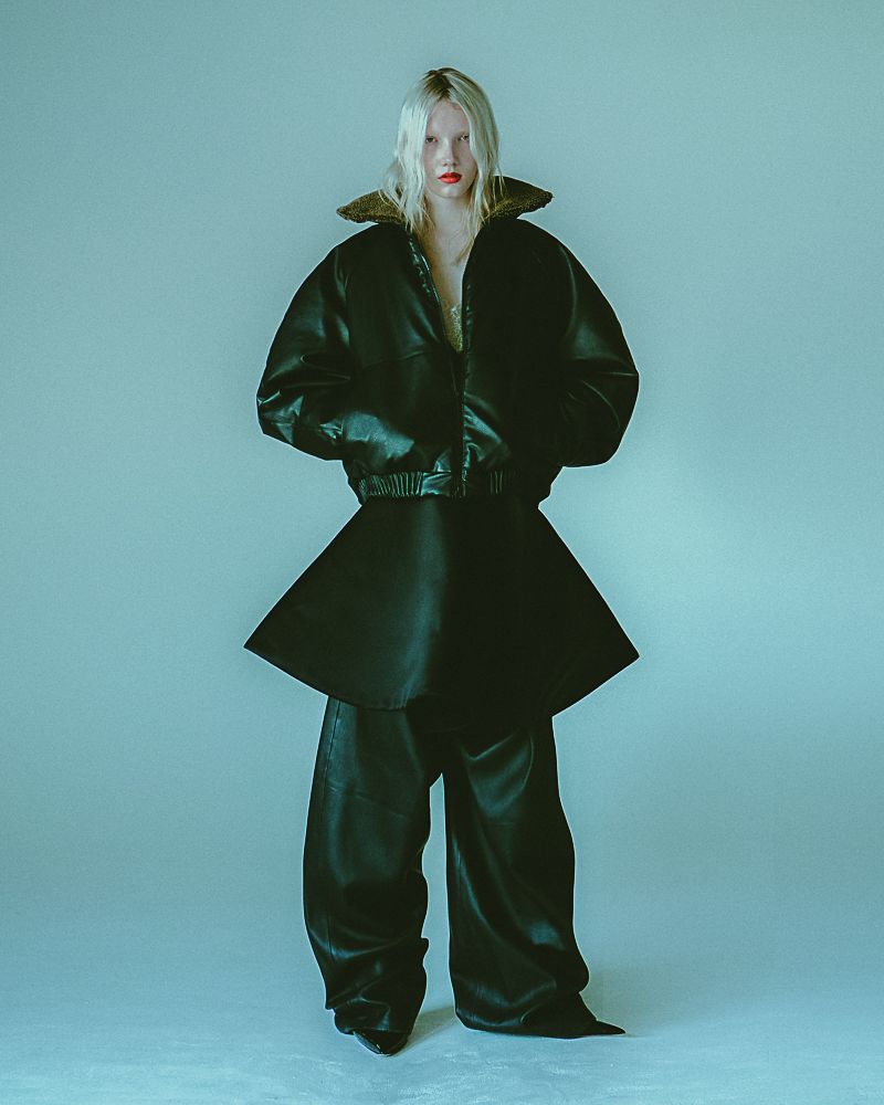 A full-length studio portrait of a tall, slender, fair-skinned model with bleached-blonde hair, wearing a black leather jacket and skirt-pants.