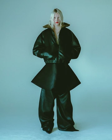 A full-length studio portrait of a tall, slender, fair-skinned model with bleached-blonde hair, wearing a black leather jacket and skirt-pants.