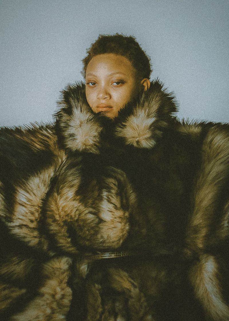 Close-up portrait of an adult woman with short hair wearing a large brown fur coat against a light blue-gray backdrop.