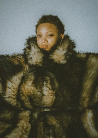 Close-up portrait of an adult woman with short hair wearing a large brown fur coat against a light blue-gray backdrop.