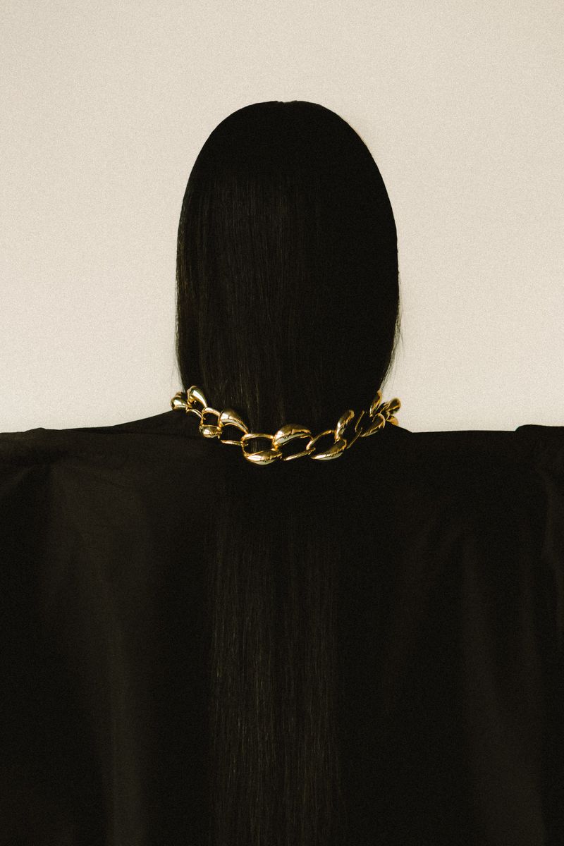 Back view of a woman with black hair wearing a gold chain necklace against a cream background, showcasing fashion and beauty.