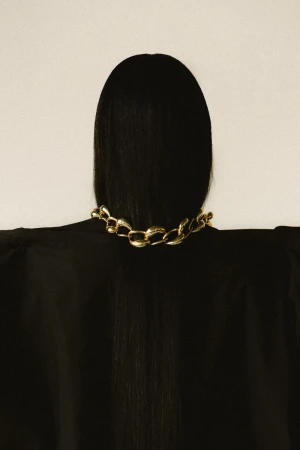 Back view of a woman with black hair wearing a gold chain necklace against a cream background, showcasing fashion and beauty.