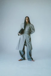 Model with long dark hair wearing a grey belted coat and trousers against a neutral background holding a clutch