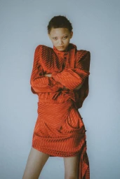 Model with short hair wearing a structured red textured dress with arms crossed against a plain grey background