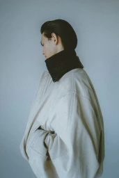 Profile view of a man with slicked-back dark hair wearing a light cream coat and dark turtleneck against a plain wall.