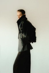 Model wearing a black leather jacket and dark trousers standing against a pale seamless studio background.