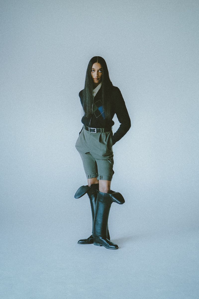 Model with long dark hair wearing a dark patterned sweater, olive shorts, and tall black boots against a plain backdrop