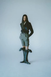 Model with long dark hair wearing a dark patterned sweater, olive shorts, and tall black boots against a plain backdrop