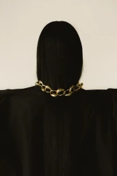 Model seen from behind with long straight black hair wearing a chunky gold necklace and black top against a neutral background.