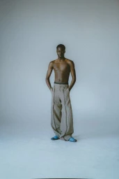 Shirtless black male model wearing wide-cut trousers against a plain grey seamless studio background.
