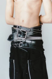Close-up of a person wearing multiple overlapping black leather belts with large buckles over black cargo pants against a plain wall.