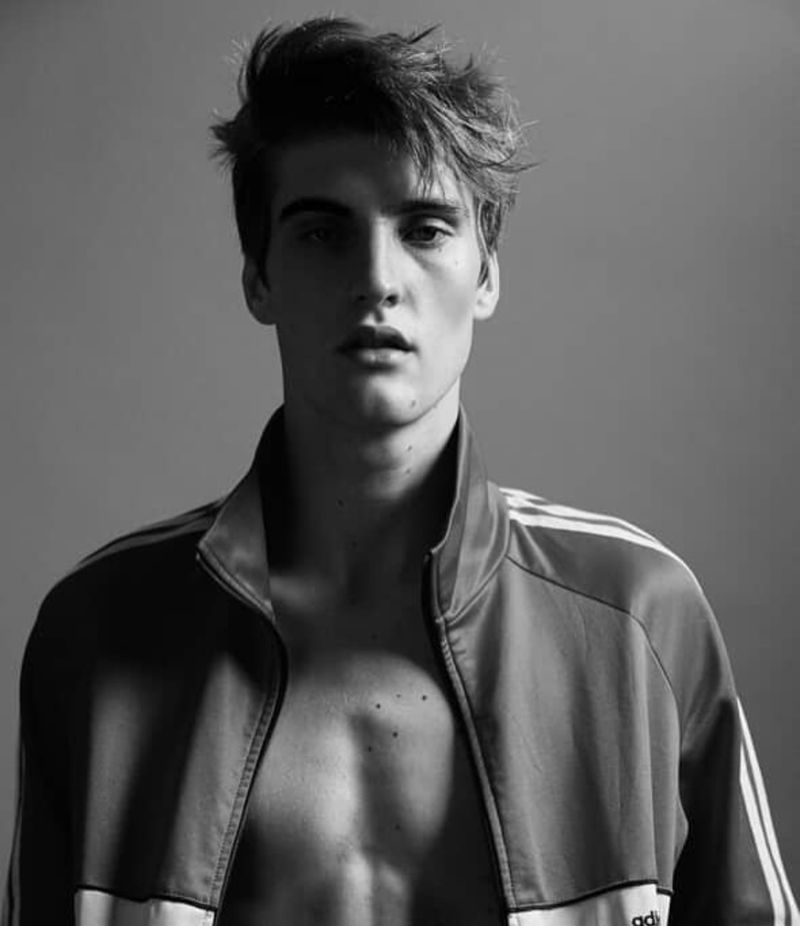 Black and white portrait of a young adult male with an open Adidas track jacket, posing in a photo studio against a plain background.