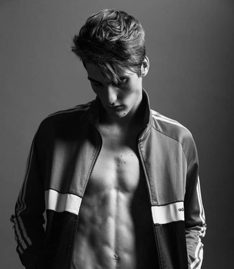 Monochrome studio portrait of a young adult man with an athletic build wearing an unzipped Adidas jacket, gazing downwards.