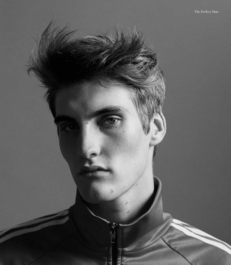 Black and white portrait of a young adult male with tousled dark hair wearing a zip-up jacket with striped sleeves against a neutral background.
