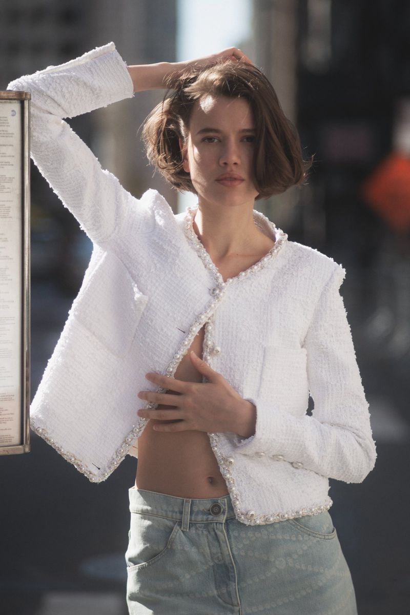 Brunette woman in white tweed jacket and light blue jeans poses outdoors against a building in an urban environment during daytime.