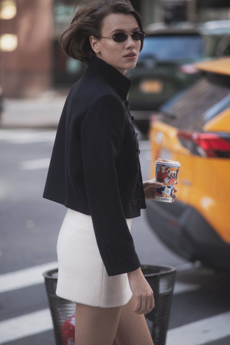 Woman in a black jacket and white miniskirt walks across a city street carrying a coffee cup with a yellow taxi in the background.