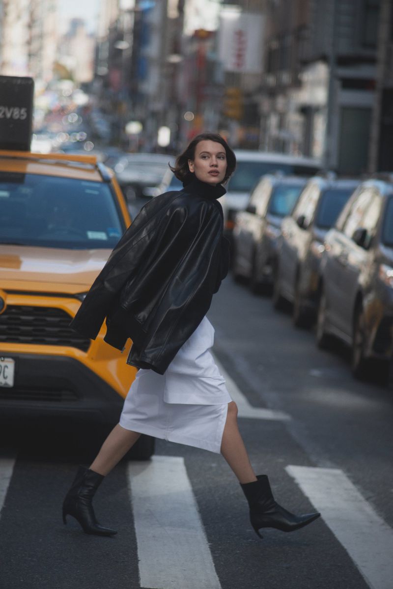 Woman in black jacket and white dress crossing urban street with taxi and cars, capturing a fashion moment in city setting.