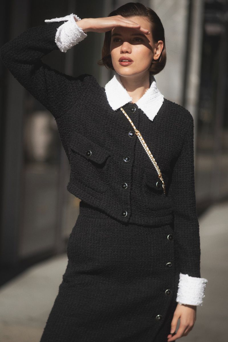 A fair-skinned adult woman in a black tweed suit and white trim is posing outdoors, shielding her eyes from the sun while looking left.