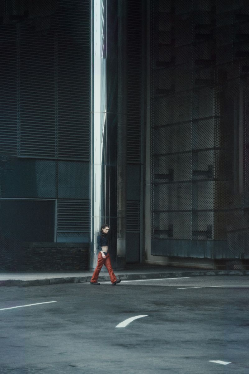 Adult wearing orange pants walking on a city street with grey buildings and a silver pillar in the background on an overcast day.
