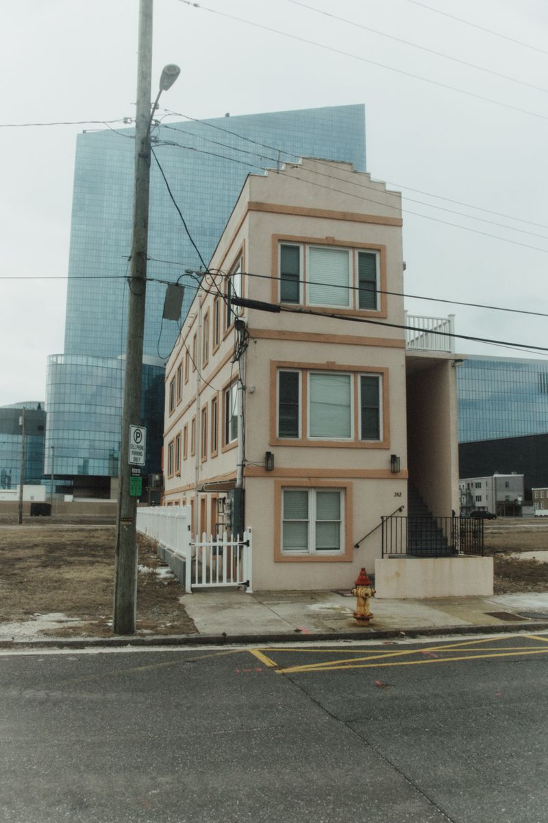 Three-story apartment building with a white fence and fire hydrant in front of a glass skyscraper on an overcast day.
