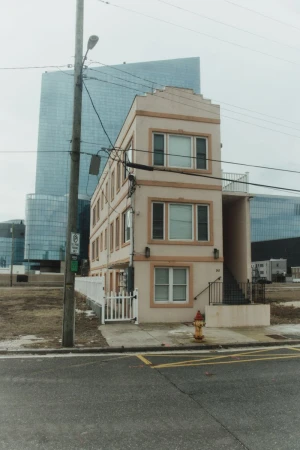 Three-story apartment building with a white fence and fire hydrant in front of a glass skyscraper on an overcast day.
