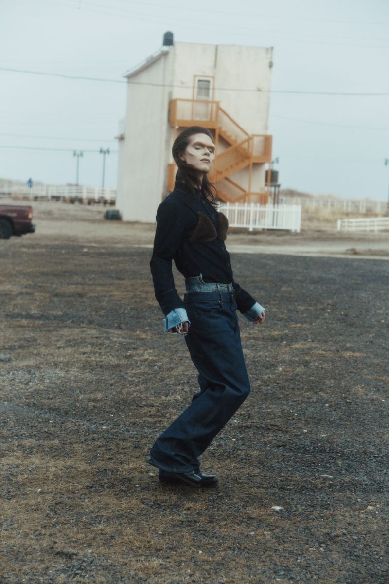 Fashionable adult woman poses in a long-sleeved black shirt and wide-legged jeans, standing outdoors on overcast day with building behind.
