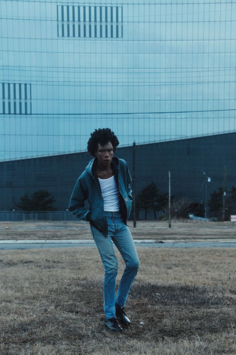 A slender African American man in jeans and jacket stands in a grassy field before a modern building with window grids and blue tones.