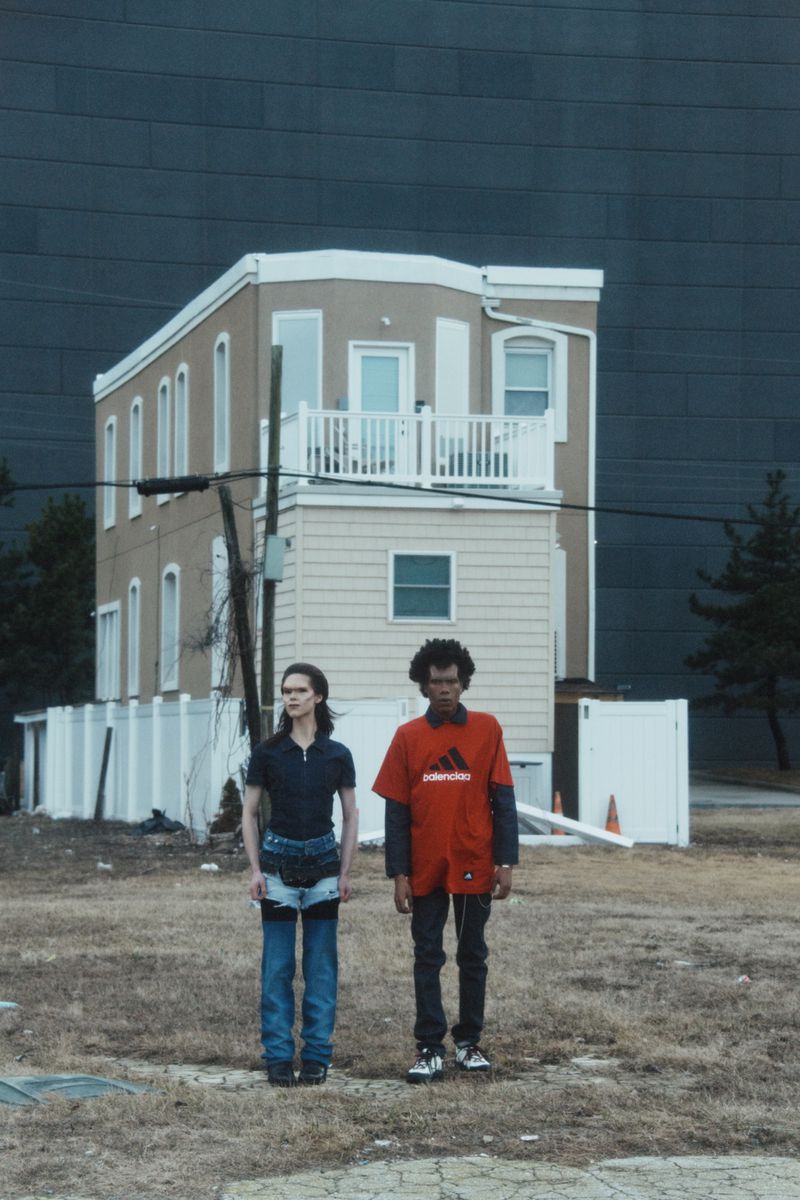 Two people stand outdoors in front of a house and a large building, showcasing streetwear fashion with a somewhat distressed aesthetic.