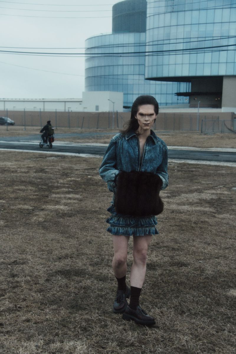 An adult with long hair and pale skin walks on patchy grass with a modern building in the background, wearing a jean jacket and dress.
