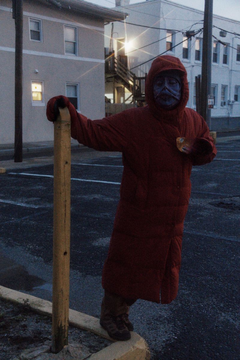 Person in a red hooded coat with painted face stands near a post in an outdoor parking lot at dusk, with buildings in background.
