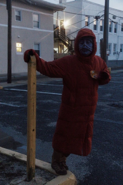 Person in a red hooded coat with painted face stands near a post in an outdoor parking lot at dusk, with buildings in background.