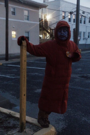 Person in a red hooded coat with painted face stands near a post in an outdoor parking lot at dusk, with buildings in background.