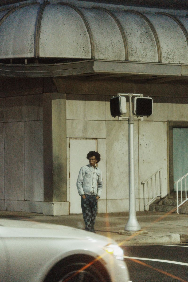 A lone person stands on a city sidewalk near a crosswalk signal, positioned in front of a building with a white car in the foreground.