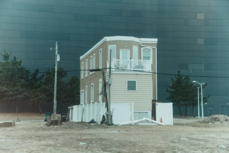Exterior view of a two-story residential building with white trim, set against a large, dark-paneled building under an overcast sky.
