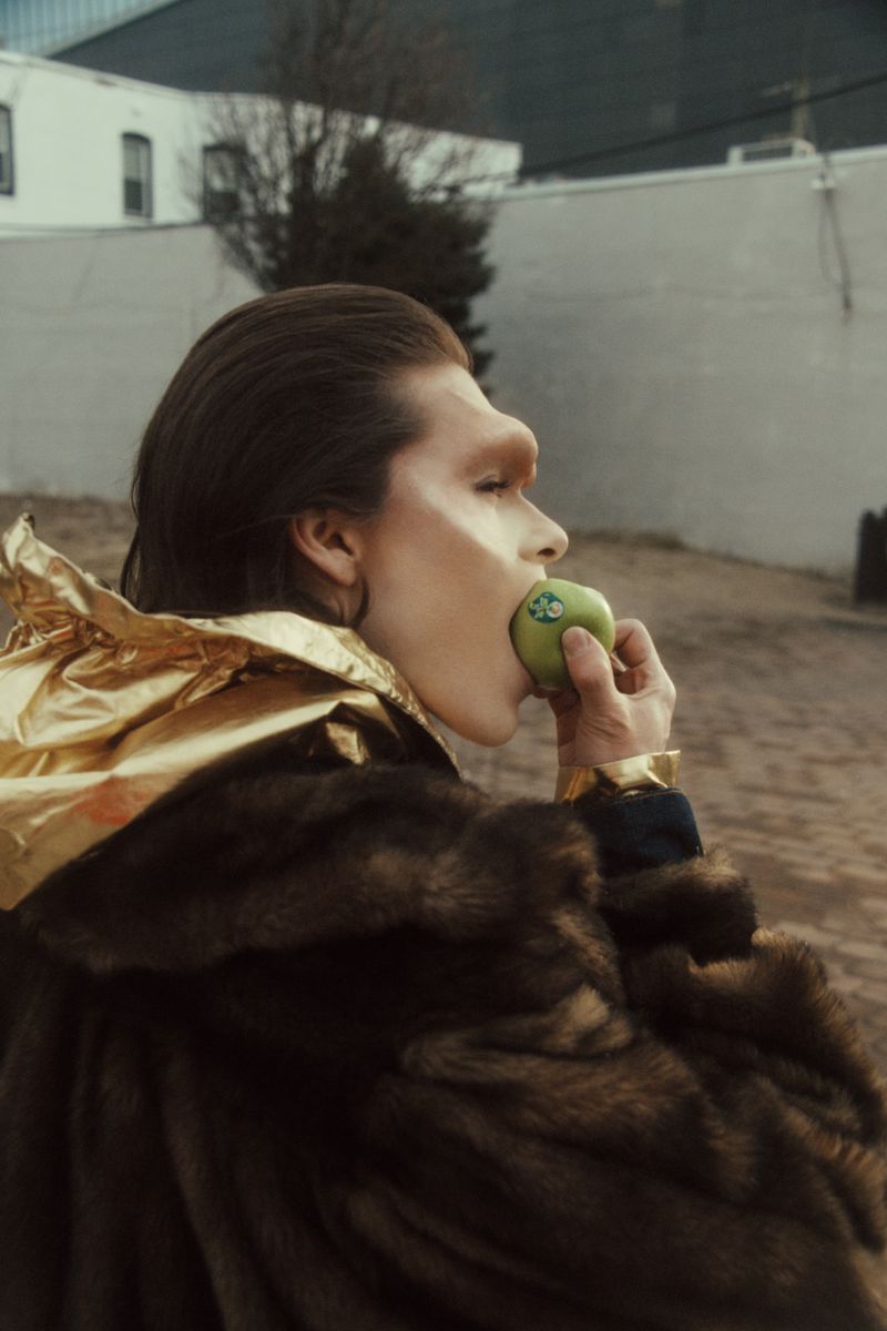 Adult is shown in profile, holding a green apple to their mouth, wearing a fur coat and gold jacket, against an urban backdrop.