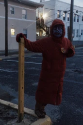 Model in a long red puffer coat leaning against a yellow post outdoors on an asphalt street with buildings behind