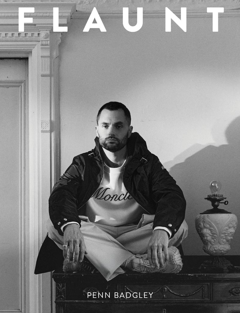 Black and white portrait of Penn Badgley for Flaunt magazine. He is seated on a table in a jacket and sweater with the brand Moncler on it.