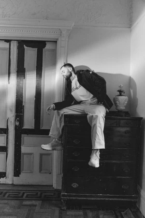 Black and white portrait of a man sitting on a dresser. The man wears a t-shirt, pants, jacket and sneakers in an indoor environment.