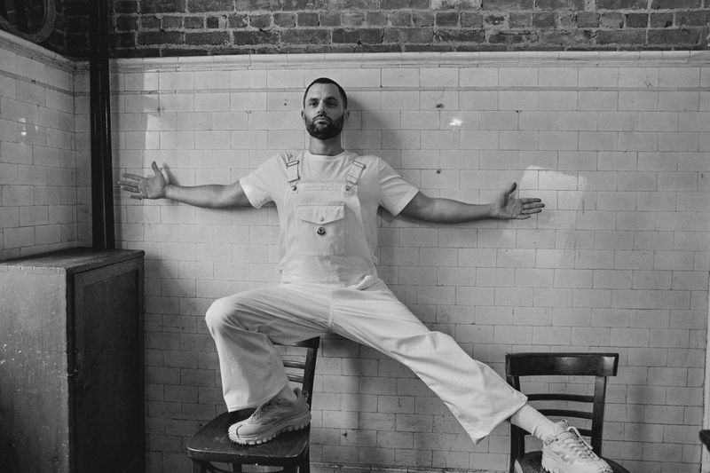 Monochrome shot of a man in white overalls balancing on chairs in a tiled room with a brick wall, arms outstretched for balance.