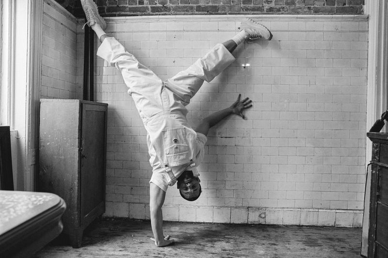 Monochrome shot of an adult male performing a handstand in an indoor tiled room with vintage furniture and window light.