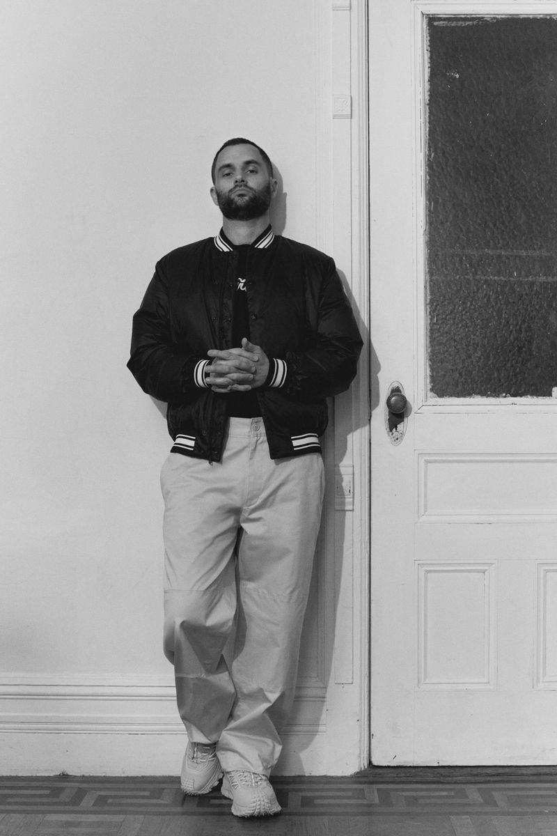 Black and white portrait of a man leaning against a white door, wearing a bomber jacket and trousers, standing indoors on a patterned floor.