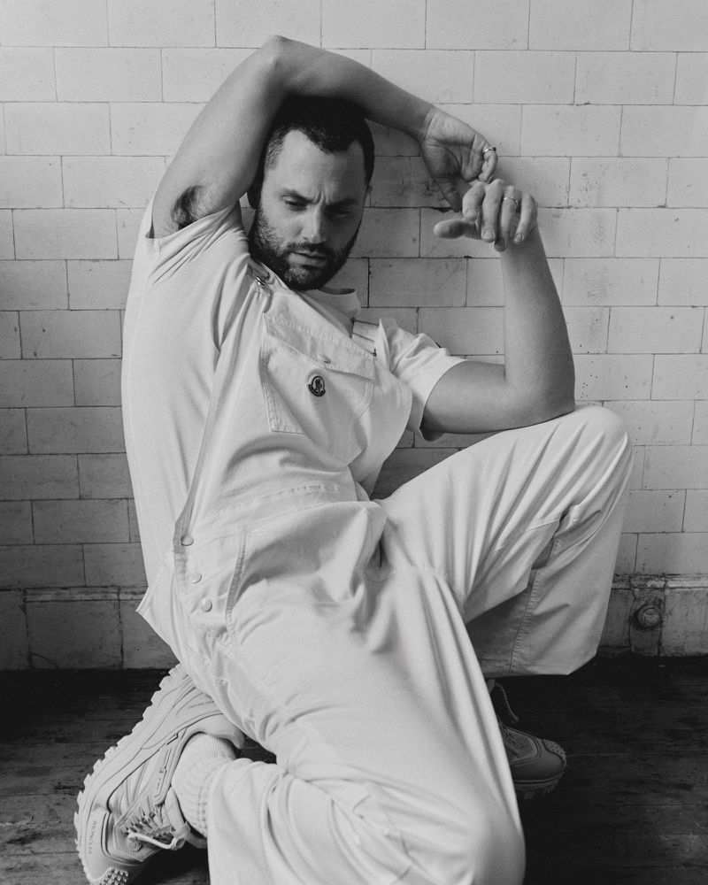 Monochrome portrait of a man in a white jumpsuit and sneakers, kneeling against a tiled wall with arms raised, looking down.