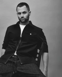 Portrait of actor Penn Badgley wearing a dark zipped jacket and white t-shirt while seated against a plain grey studio backdrop.
