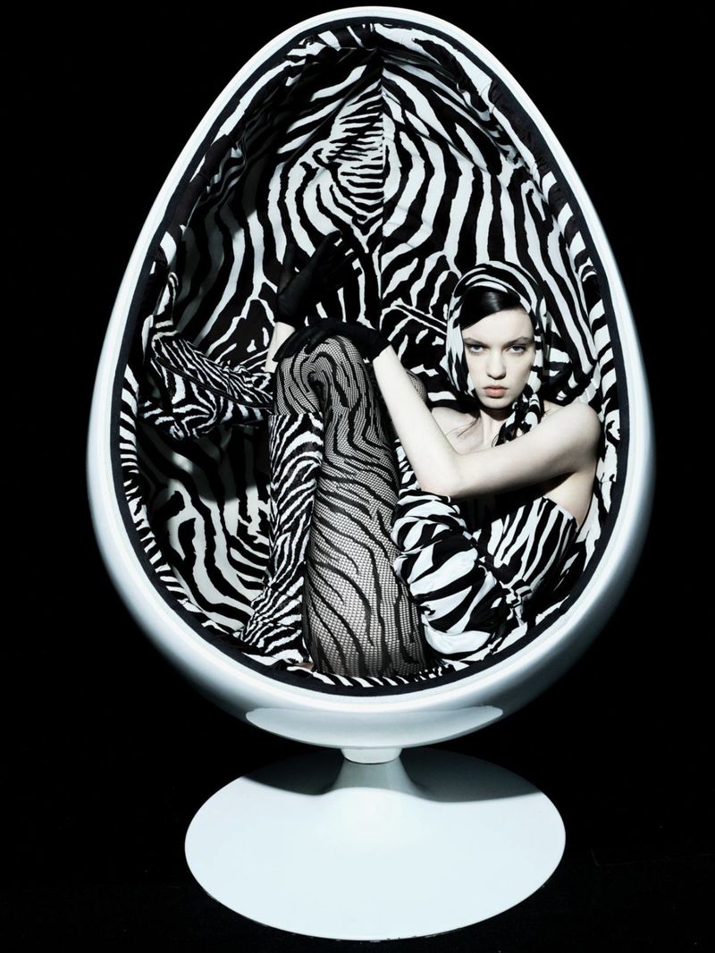 A woman wearing zebra-striped clothing sits in a white, egg-shaped chair with zebra-striped fabric against a black background in a fashion shot.