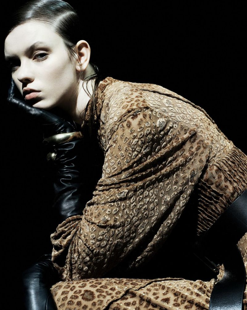Elegant woman in patterned dress with black leather gloves posing against dark backdrop with studio lighting for a fashion editorial shot.