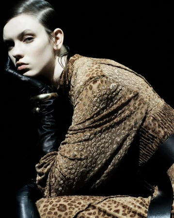 Elegant woman in patterned dress with black leather gloves posing against dark backdrop with studio lighting for a fashion editorial shot.