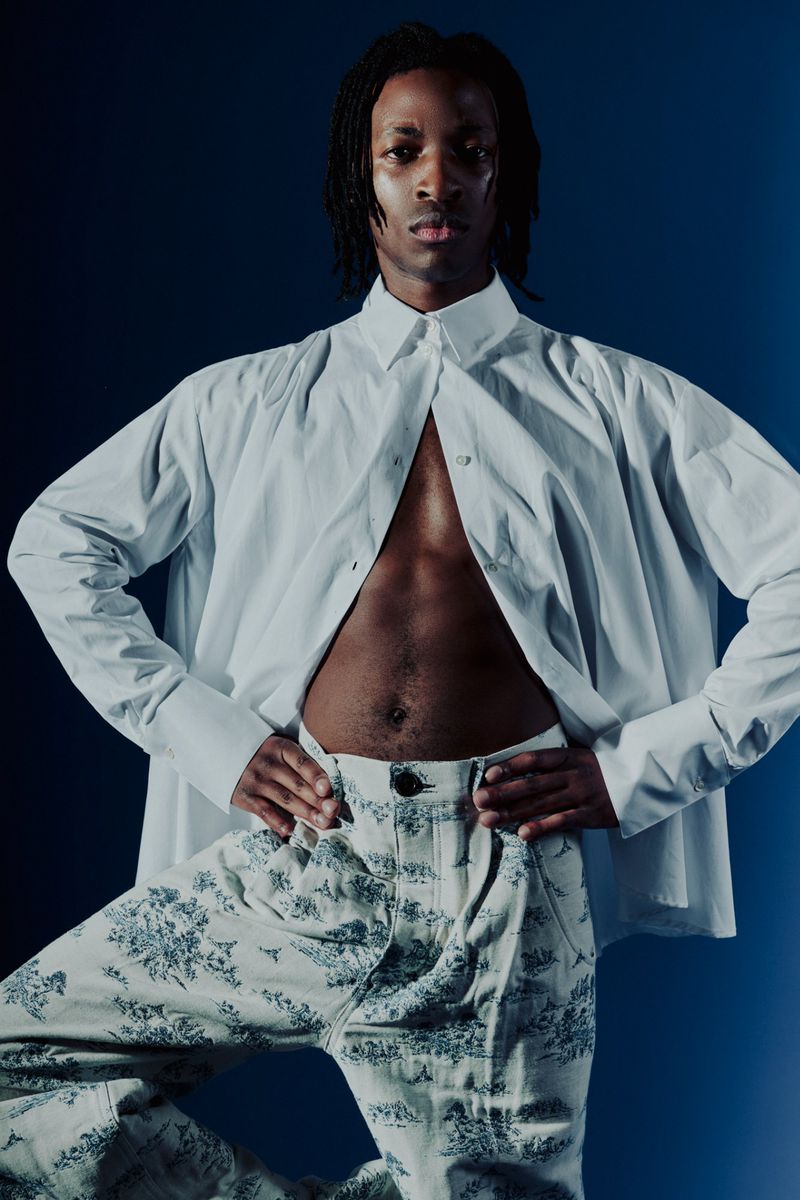 An adult Black male poses in a white shirt and patterned pants against a dark blue gradient background in a fashion photography studio.