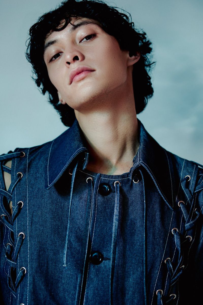 Portrait of a person wearing a denim jacket with decorative lacing, posing against a blurred, light-colored background in a studio setting.