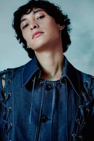 Portrait of a person wearing a denim jacket with decorative lacing, posing against a blurred, light-colored background in a studio setting.